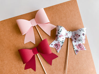 Set of 12 3D paper bow cupcake toppers in pink, dark red, and floral rose pattern. All are affixed to a food safe pick for decoration cakes or cupcakes.