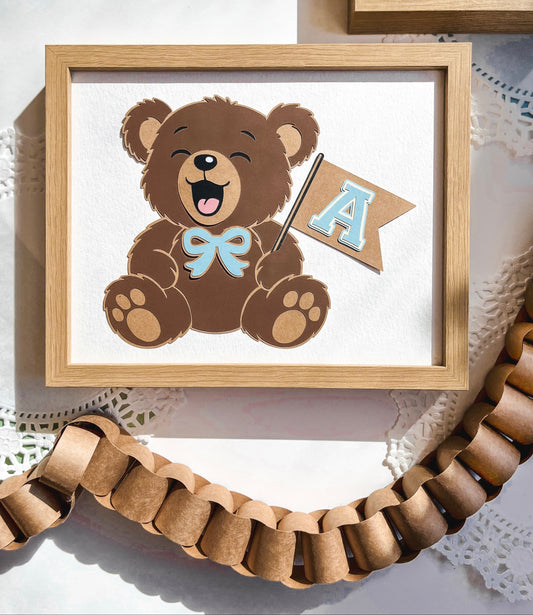 Framed picture of a teddy bear with a bow tie and a small flag with 'A' on a white background. Customizabe to any letter and in the colors pink, blue, and green! 